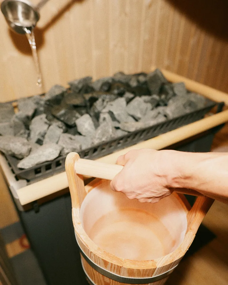 Eine Hand hebt den Aufgusskübel vor die Saunasteine, aus der Kelle läuft Wasser